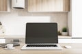 Remote working concept with blank black laptop screen in cozy kitchen interior with light furniture, black diary and mug of coffee