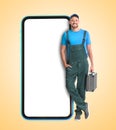 Repair service - just call. Happy professional repairman holding toolbox and smartphone with blank screen on beige background,