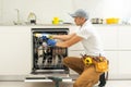 Repairman in uniform repairs dishwasher in kitchen. Young man specialist unscrews parts with screwdriver checking state Royalty Free Stock Photo