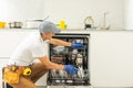 Repairman in uniform repairs dishwasher in kitchen. Young man specialist unscrews parts with screwdriver checking state Royalty Free Stock Photo