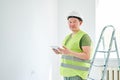 Repairs in the apartment, a man in a green vest and a white helmet is in a white room with a tablet in his hands