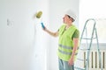 Repairs in the apartment, a man in a green vest and a white helmet stands in a white room with a roller in his hands