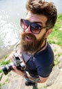 Reporter photographer concept. Man with beard and mustache wears sunglasses, water surface on background. Guy shooting