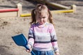 A resent little girl is standing in a sandbox holding a plastic shovel