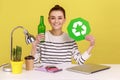 Woman holding green recycling sign in hand and glass bottle, sitting on workplace with laptop. Royalty Free Stock Photo