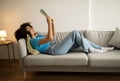 Rest, relax and study at home. Glad young black curly woman reading book, lying on sofa, enjoy spare time Royalty Free Stock Photo