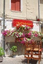 building in an Italian town, red wooden shutter, restaurant tables with chairs, colorful flowers fixed on the wall