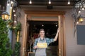 Young waitress in grey apron standing at the entrance, holding open sign, smiling Royalty Free Stock Photo