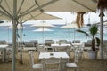 Restaurant tables and chairs setup under umbrella in white color and long chair in blue on Ornos sand beach with seaview