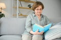 Restful reading. Portrait of thoughtful aged woman reading favorite literature at cozy home. She is lying on pillows on Royalty Free Stock Photo