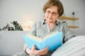 Restful reading. Portrait of thoughtful aged woman reading favorite literature at cozy home. She is lying on pillows on Royalty Free Stock Photo