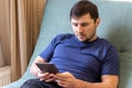 A resting man in a blue t-shirt sits on a green sofa with an e-book in his hands. Gray e-reader electronic book or tablet