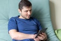 A resting man in a blue t-shirt sits on a green sofa with an e-book in one hand. Gray e-reader electronic book in male hands