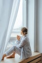 Calm girl in pajamas with cup of tea or coffee sitting and drinking on the window-sill at home. Side view Royalty Free Stock Photo