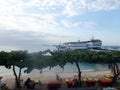 Resting tourists are sitting at the tables at the sea beach restaurant under the trees. Large ships in the background