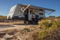 A retired Australian woman sits under the awning of a modern caravan adjacent to a beach in a bright clear day in Australia