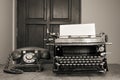 Retro aged black typewriter with paper blank, telephone on wooden table. Vintage style sepia photo