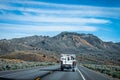 Retro camper mounted on pickup truck with smoke blowing out the back drives down two lane highway through sagebrush to badlands