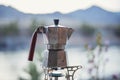 A retro coffee maker made of aluminum stands on a gas burner against the backdrop of mountain peaks on a hike. A tourist