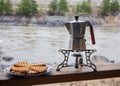 A retro coffee maker made of aluminum stands on a gas burner against the backdrop of a river bank on a hike.