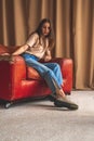 Retro fashion. Teenager girl in classic denim jeans and urban sneakers sitting on old luxury leather chair in studio Royalty Free Stock Photo