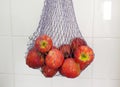 Reusable thread mesh filled with red apples hanging on the ceramic tiles wall. Storage for future use