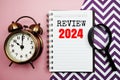 Review 2024 text on paper notebook with alarm clock top view on pink background