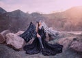 Revived gargoyle, queen of night watching sunrise, girl in long light black dress with black feather wings sits on rocks