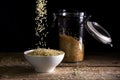 Rice grains falling into a white bowl beside a glass jar with ri