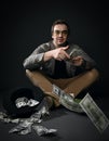 Rich insolent adult man in jacket, jeans and shoes sits on floor by upside down hat full of dollars throwing cash Royalty Free Stock Photo