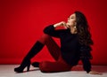 Rich romantic adult woman with long curly brunette hair wearing black pullover, red pants and high boots sits on floor Royalty Free Stock Photo