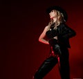 Rich sexy curly blonde woman in black leather pants, top and wide-brimmed hat stands sideways looking at copy space
