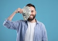 Rich young guy closing his eye with fan of money, happy over big success, blue studio background