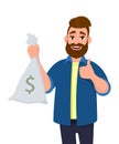 Rich young man is holding money, cash bag in hand and gesturing thumbs up sign. Successful business and finance concept. Royalty Free Stock Photo