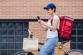 Rider woman wearing red backpack delivering food on a bike, checking order with smart phone while standing on street in city. Royalty Free Stock Photo