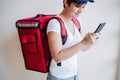 Rider woman wearing red backpack delivering food, checking order with smart phone while standing on street in city. Delivery