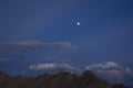 Ridge of gray brown desert mountains under a dark blue evening sky with gray clouds and a full moon with stars Royalty Free Stock Photo