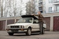 A man folding the roof of white classic convertible BMW E30 car. Old garages and modern building in the background.