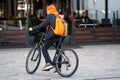 A young man in a brightly colored hat and with a backpack on a bicycle in a paved area near a cafe