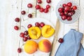 Ripe colorful peaches halved and whole, sweet cherries on white plate, crystal bowl, knife, plank wood table, blue napkin, styled