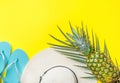 Ripe pineapple on green palm leaf white straw hat blue slippers on bright yellow solid background. Summer vacation fun