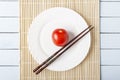 Ripe red tomato and chopsticks on white plate. Top view on wooden table with bamboo mat. Vegetarian food concept Royalty Free Stock Photo