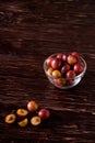 Ripe sweet plum fruits with water drops in glass bowl near to scattered sliced half plums on dark moody wood table background