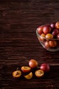 Ripe sweet plum fruits with water drops in glass bowl near to scattered sliced half plums on dark moody wood table background