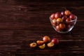 Ripe sweet plum fruits with water drops in glass bowl near to scattered sliced half plums on dark moody wood table background