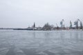 A river with floating melting pieces of ice. There are many construction cranes and tower on the shore.