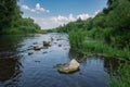 A river with groups of scattered stones running to the horizon. Lush green banks, blue sky with puffy white clouds, water surface Royalty Free Stock Photo