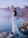 A river nymph in a white lace dress stands on a rock by the lake. The princess has a beautiful wreath with seashells