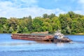 A river tug is pushing a rusty barge along the river along the shore