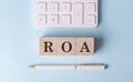 ROA word on wooden block with pen and calculator on blue background